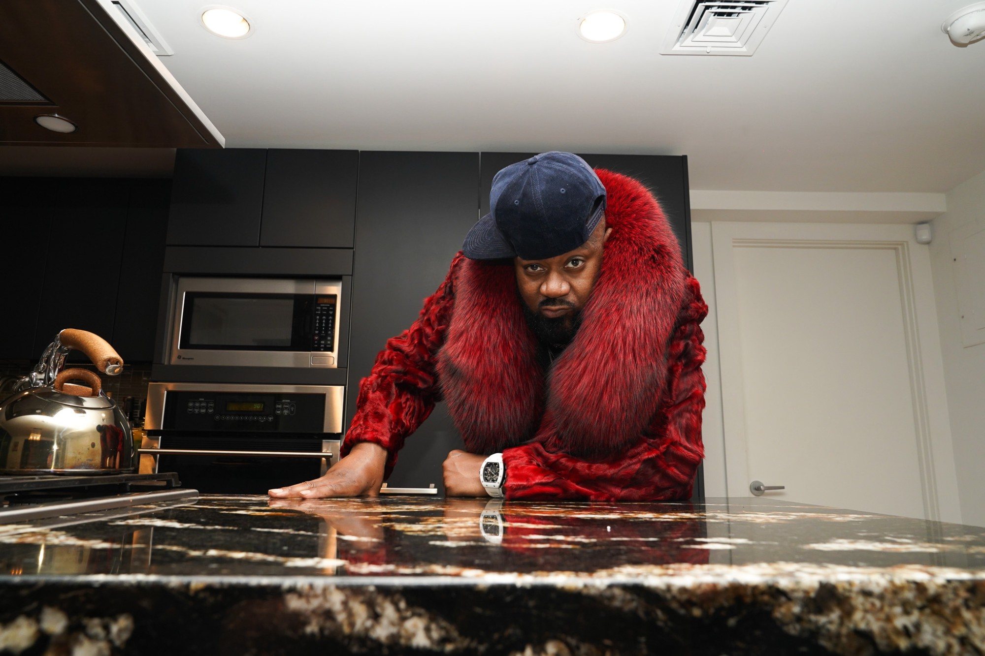 Ghostface Killah in a red fur coat leaning over a granite kitchen counter, looking into the lens
