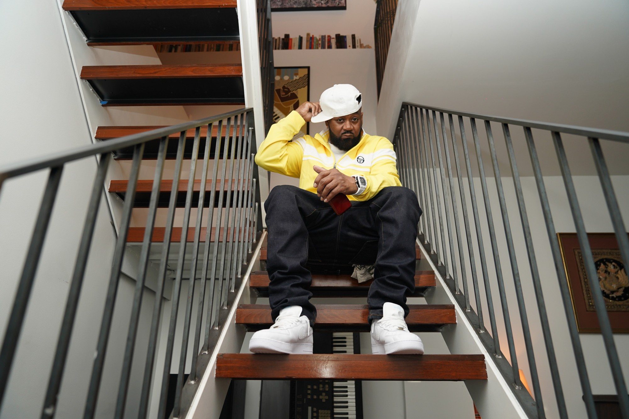 Ghostface Killah on an open staircase in a yellow track jacket, touching the brim of a white cap